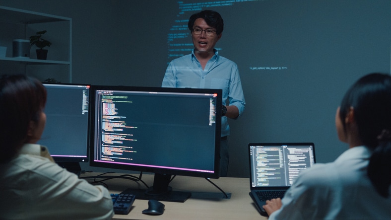 Young Asian man software developers mentor leader manager talking to executive team analyzing source code in office at night. Programmer development.